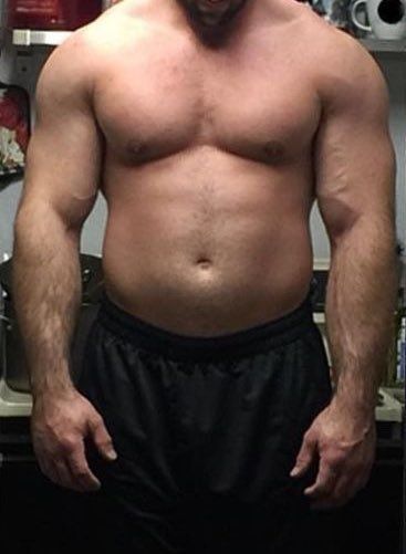 the image shows a shirtless, muscular man with a slightly overweight physique. he's standing in what appears to be a home kitchen, wearing dark shorts. his body features welldeveloped biceps, triceps, chest, and abdominal muscles. the man seems to be middleaged and has visible body hair. the background indicates a kitchen setting with shelves and some food items visible. the overall impression is of a strong, athletic man who may be involved in weight training or bodybuilding.