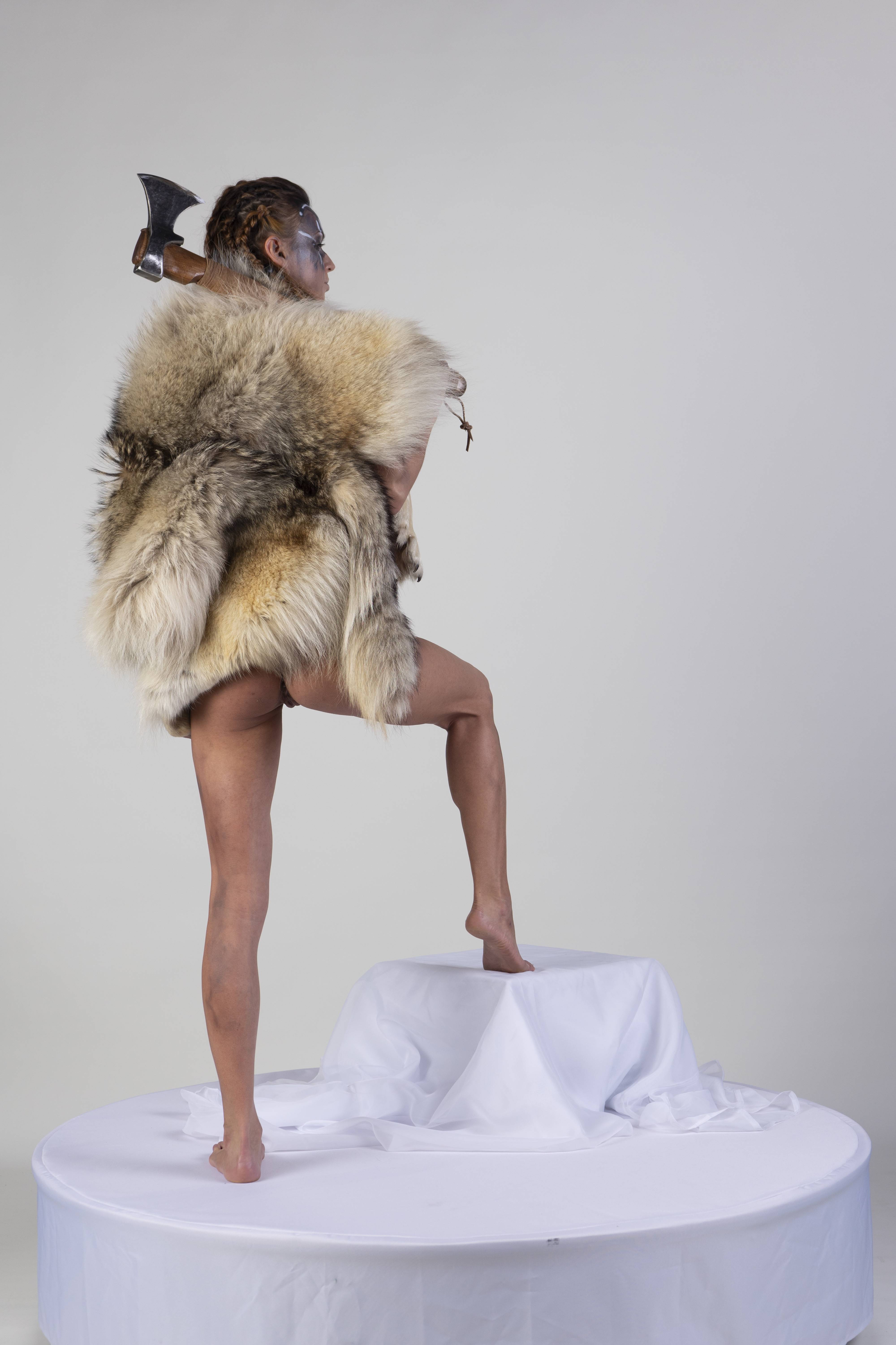 a barefoot woman in a fur coat stands on a white pedestal with an axe over her shoulder. the woman is wearing face paint and looking to the right. the background is a plain white.