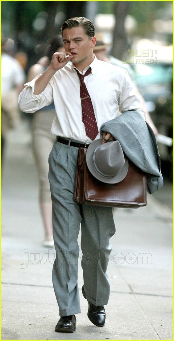 a man in a classic suit and tie walks down a city street carrying a briefcase and smoking a cigarette. he is wearing a gray suit with a white shirt and a dark tie. he is also wearing a hat. the man is walking in the street which is empty. he is walking with a cool, vintage style. he has a suave gentlemanly look.