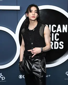 a photo of sunghoon from the kpop group enhypen at the american music awards. he is dressed in a stylish black outfit consisting of leather pants, a mesh top, and various accessories. his long hair is styled loosely, and he is posing for a photo on the red carpet. the background features the american music awards logo.