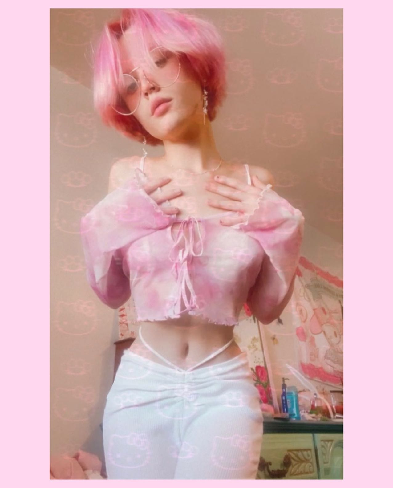 a young woman with short, bright pink hair is posing for a selfie. she is wearing a pink tiedye crop top and highwaisted white pants.  the outfit is pastel and girly.  she's wearing round glasses. the background is a blurry bedroom setting, with a dresser and other elements visible. the overall style is heavily influenced by current social media trends.  the image has a filtered, soft, almost dreamy aesthetic.