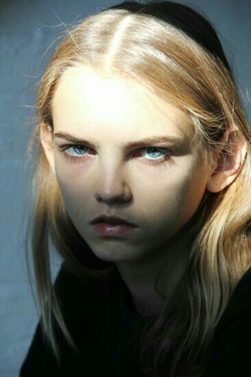 a portrait of a young woman with blonde hair and striking blue eyes. she is facing the viewer with a serious expression and an intense gaze. the lighting creates strong contrasts of light and shadow on her face, highlighting her features. she is wearing a black top and the background appears to be a muted blue.