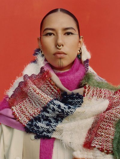 a portrait of a woman with a nose ring and traditional facial tattoos. she is wearing a colorful patterned scarf and earrings. the background is a solid red color.