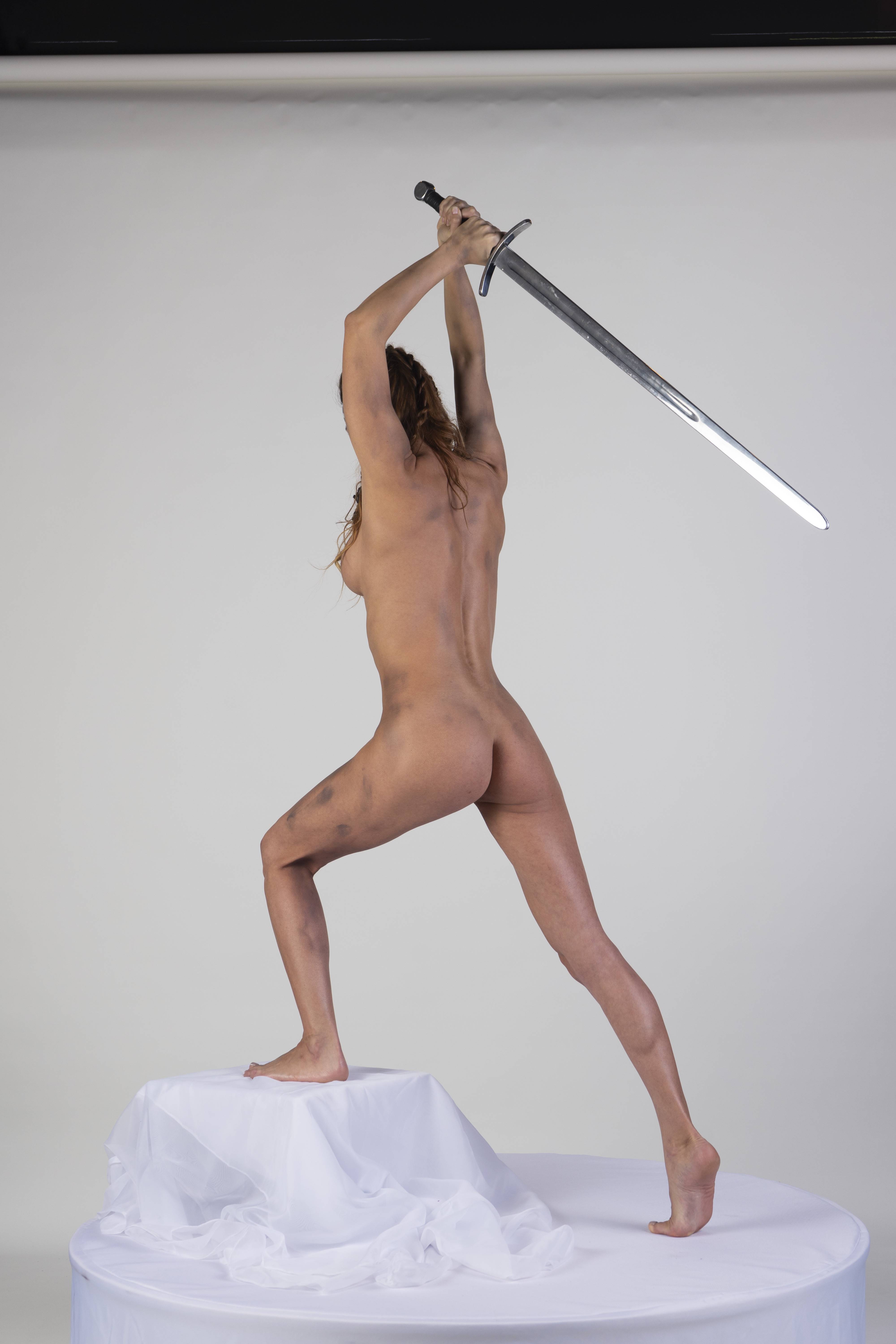 a nude woman stands with her back to the viewer, holding a sword above her head in a studio setting. she is standing on a white, round platform covered with a white cloth. the background is a solid white. the woman appears to be in a dynamic pose, possibly striking with the sword.