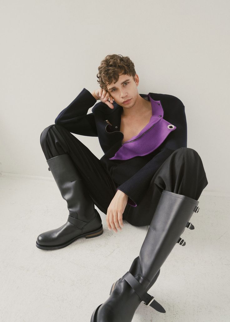 a man in a black jacket with a purple lining and black pants, sits on a white backdrop. the man is wearing black leather boots and has his hand resting on his face.