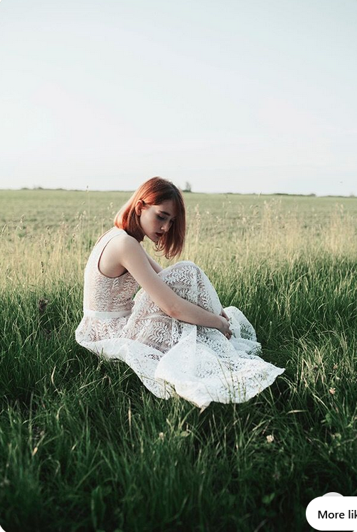 a young woman with red hair is sitting in a field of tall grass. she is wearing a white lace dress and is looking down at her lap. the background is a bright, overcast sky. the overall feeling of the image is one of peace and serenity.