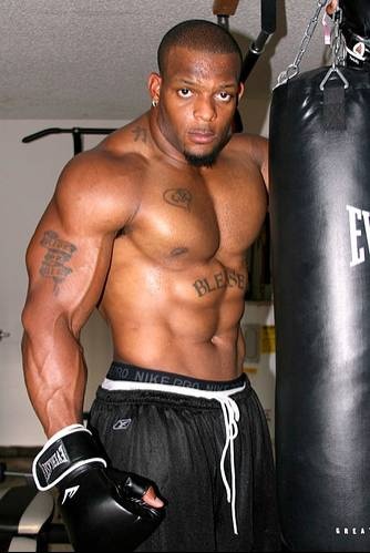 a shirtless muscular man with tattoos stands in front of a black boxing bag. he's wearing black athletic shorts and boxing gloves. he looks focused and determined, ready to train.