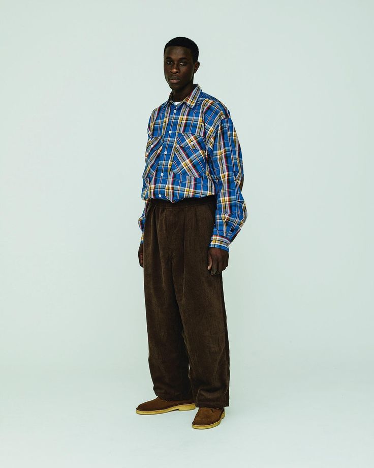 the image shows a darkskinned man standing against a light teal background. he's wearing a blue and yellow plaid longsleeved shirt, loosefitting brown corduroy pants, and brown loafers.  his pose is relaxed and straightforward. the overall style is casual and somewhat retro. the lighting and background are simple and clean, drawing attention to the clothing.