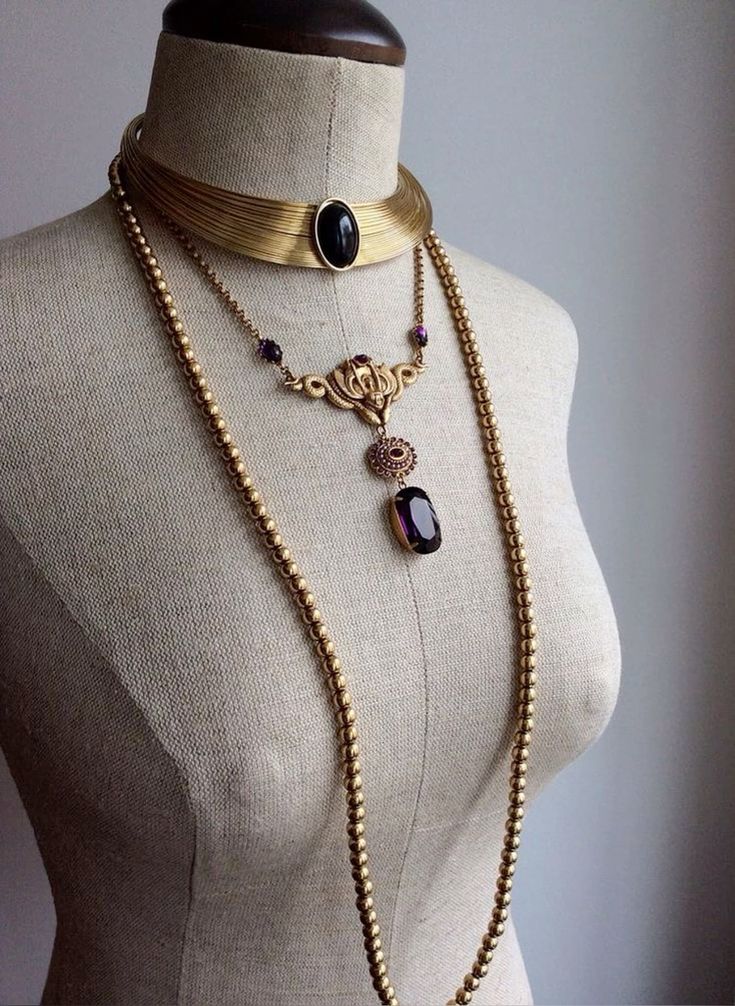 a mannequin wearing a gold layered necklace set. the set includes a choker with a black stone, a pendant necklace with a purple stone, and a long chain necklace.