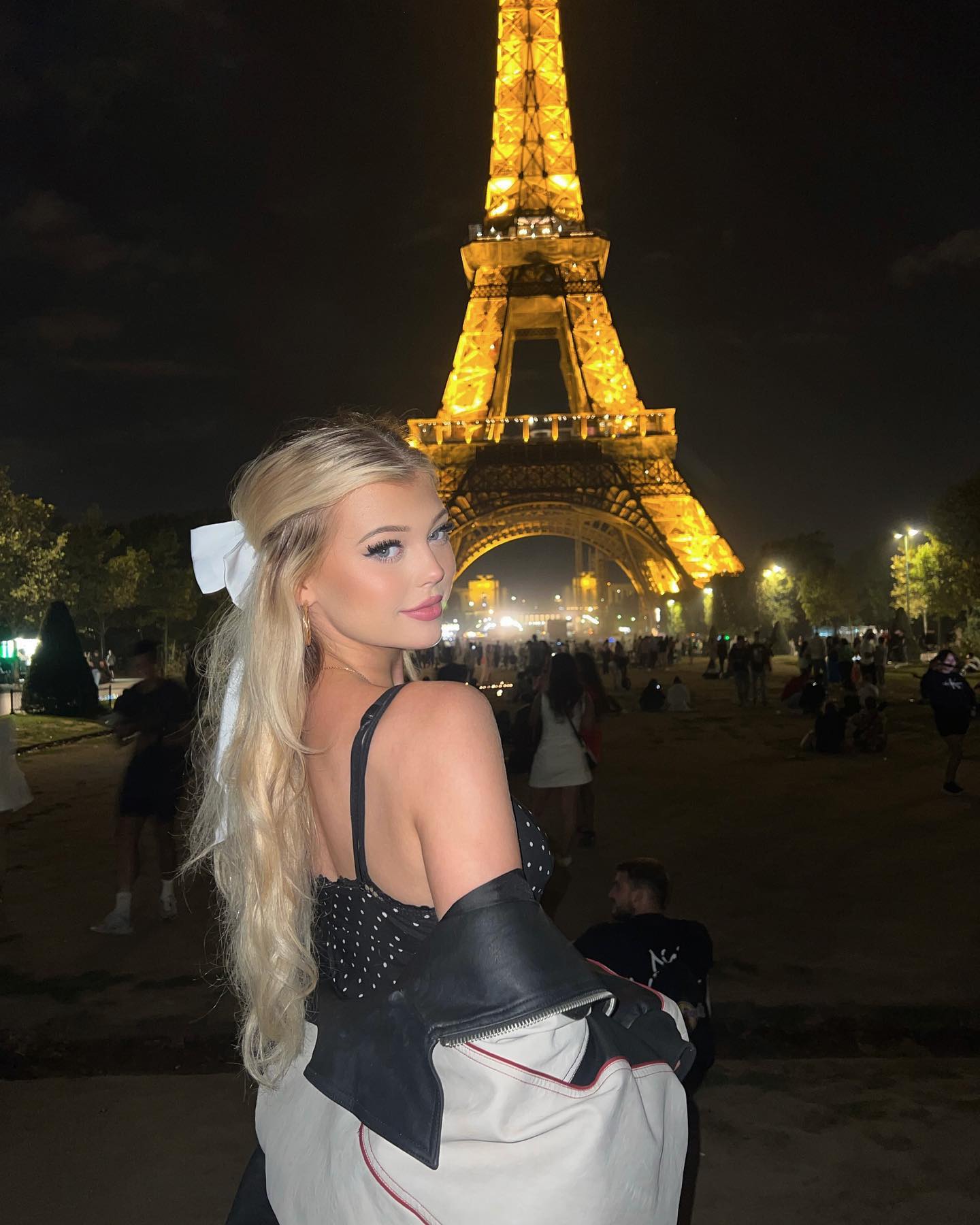 a woman with blonde hair stands in front of the eiffel tower in paris at night. she is wearing a polka dot top and a leather jacket. the eiffel tower is lit up in the background. there are other people walking around in the background.