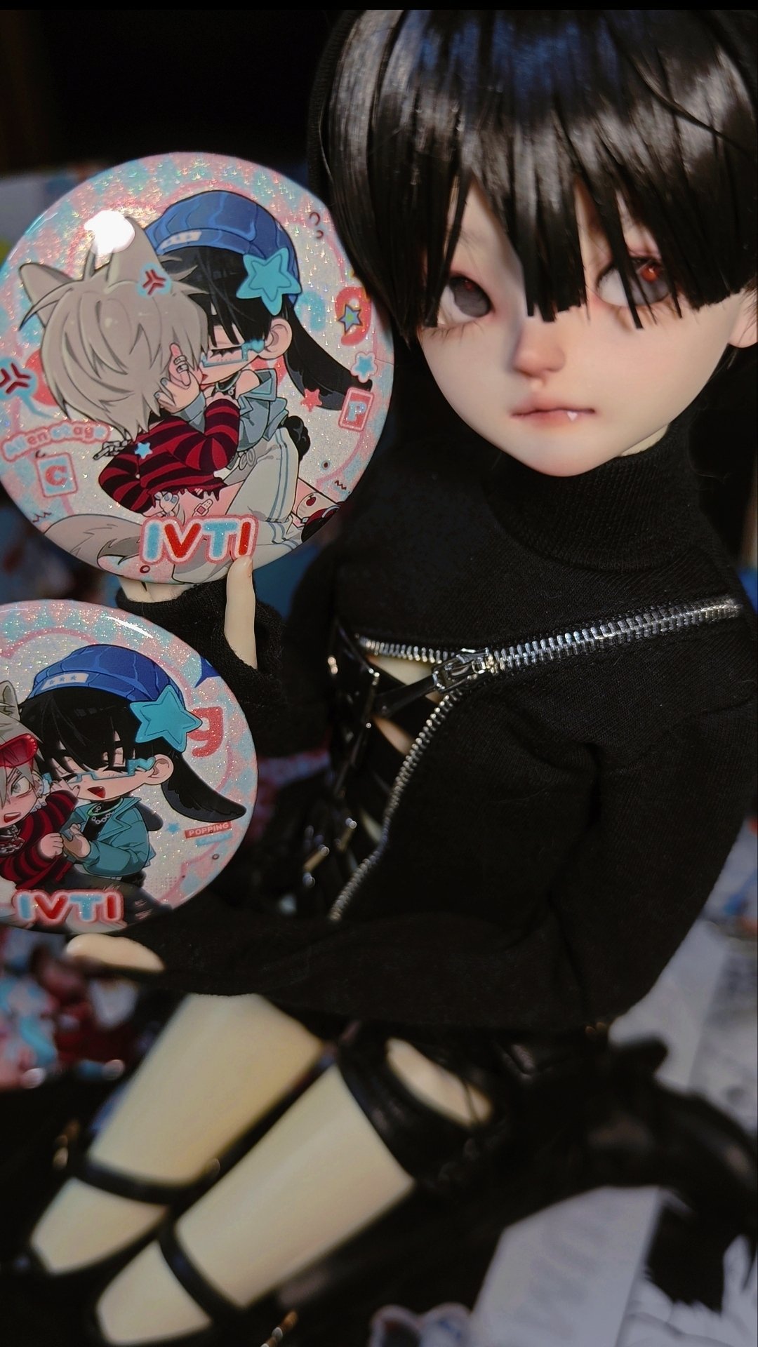 a doll is holding up two different button pins featuring a cute anime couple. the doll has black hair and is wearing a black shirt with a zipper and straps, and black boots. they are holding the buttons up in front of their face and looking off to the side. the doll has a cute and innocent expression.