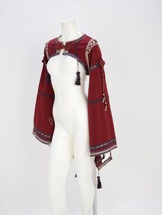 a red cropped jacket with bell sleeves and embroidery is displayed on a white mannequin. the jacket features tassels and a bohemianinspired design.