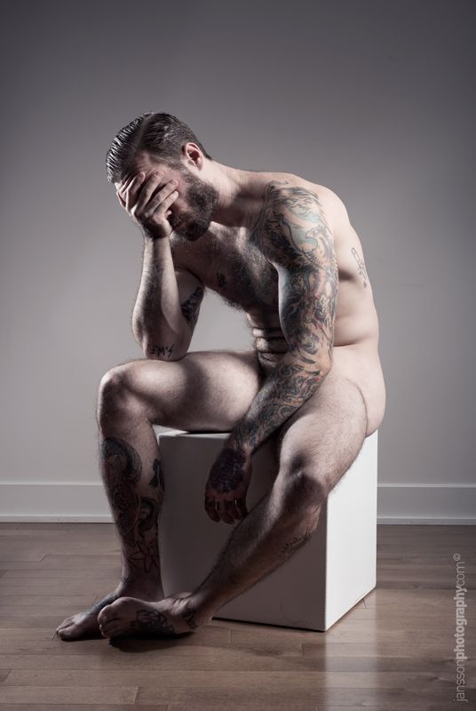 a man sits on a white cube with his face buried in his hands.  his body is covered in tattoos. he is looking down with a sad, thoughtful expression. the background is a light gray wall. the floor is wood. the image is in black and white.