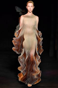 a woman in a beautiful, flowing gown on a runway. the gown is made of a translucent fabric with intricate folds and pleats, and the woman's pose highlights the graceful movement of the fabric. her makeup and hair are simple and elegant, allowing the dress to be the focus of the image.