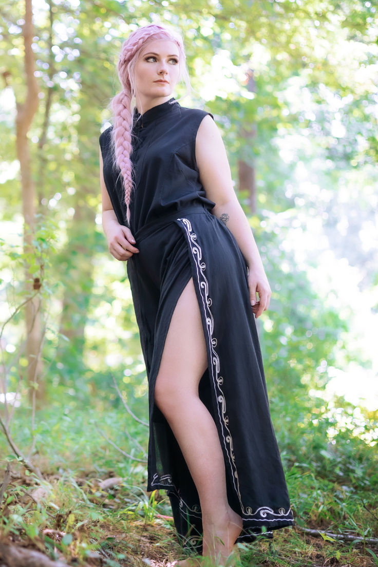 a young woman with long pink braided hair stands in a forest wearing a black maxi dress with embroidered side detailing.  the dress has a side slit revealing her leg. she is barefoot and looking off to the side, her pose suggesting a sense of calm and serenity. the setting is a lush, green forest with dappled sunlight. the overall aesthetic is bohemian and ethereal.