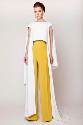 a woman wearing a white top and long yellow pants with a long white cape is posing for a fashion photograph. the background is white and the image has a very soft and elegant aesthetic.