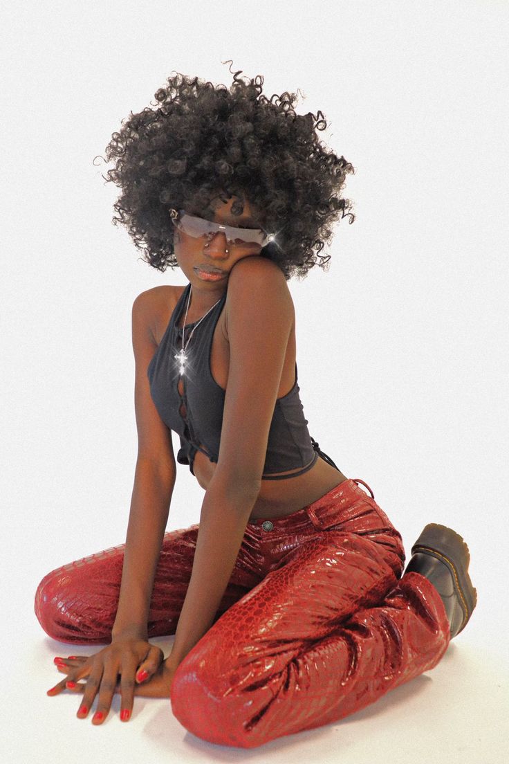 a woman with a large afro and sunglasses sits in a pose on the floor. she wears red, snake print pants, and a black cropped top. she looks directly at the camera.