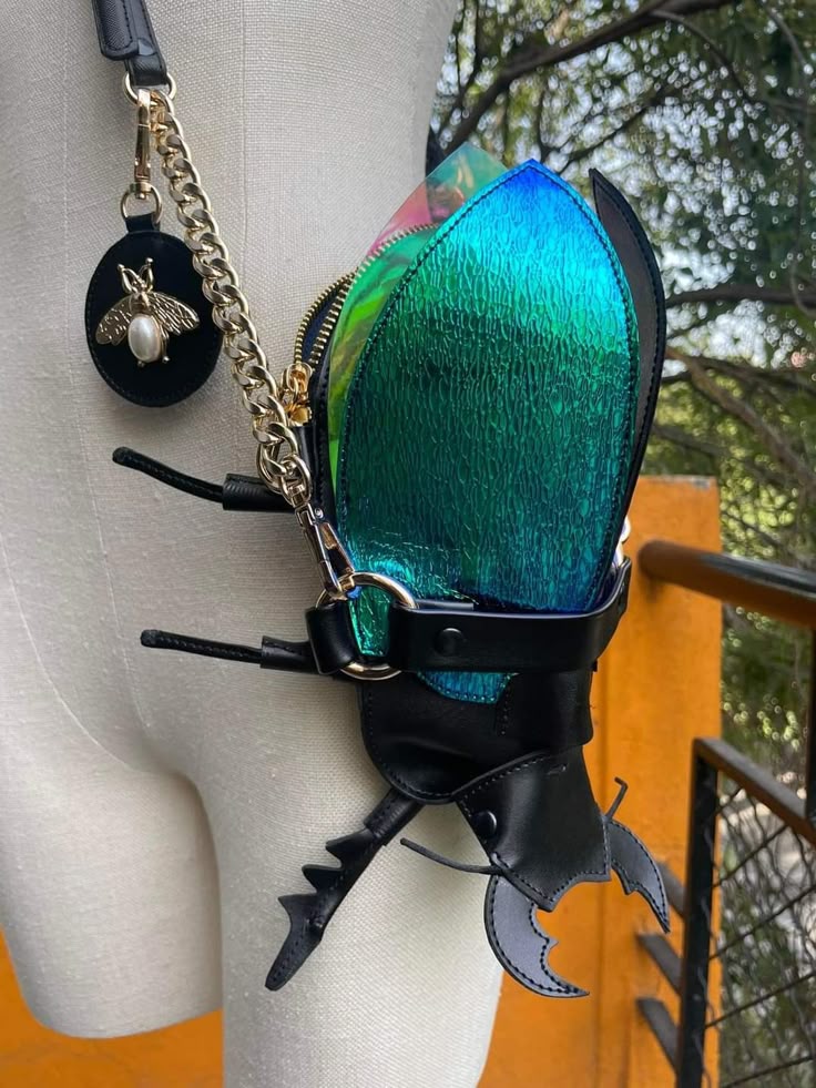 a whimsical and unusual beetleshaped handbag is displayed on a mannequin. the bag features iridescent wings that shift in color from blue to green, mimicking the natural shimmer of insects. these wings are enclosed by black leather accents that add structure and contrast. additional black leather details form the beetle's legs and mandibles, completing the insectile design. a gold chain and black leather strap allow the bag to be worn as a shoulder bag. a small, round black charm adorned with a gold bee and pearl detail hangs from the chain, adding a touch of elegance and charm.