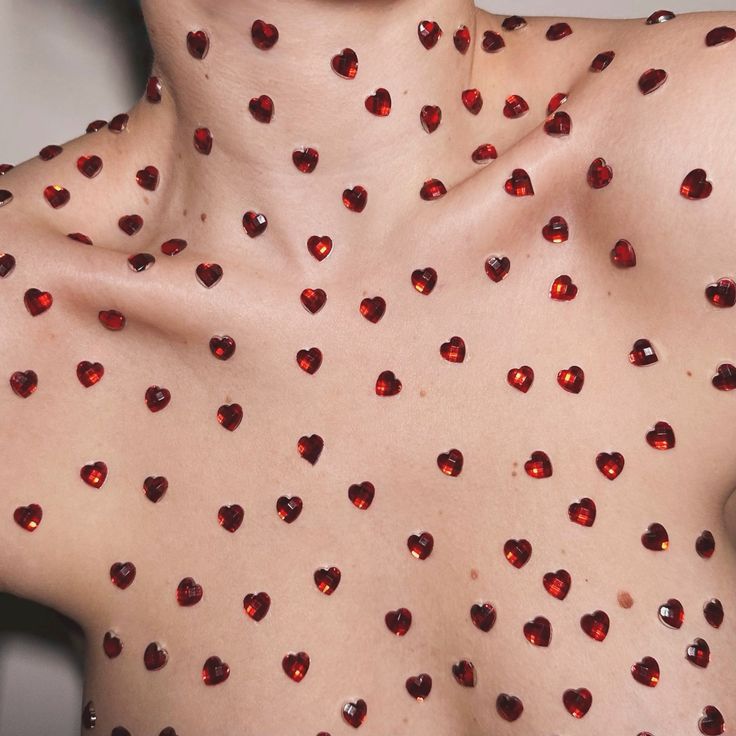 a closeup image shows a person's upper chest and neck adorned with numerous small, dark red, heartshaped rhinestones. the rhinestones are evenly spaced, creating a repeating pattern across the skin. the skin is lighttoned, and the overall aesthetic is romantic and visually appealing.  the image's focus is on the pattern and texture created by the rhinestones.