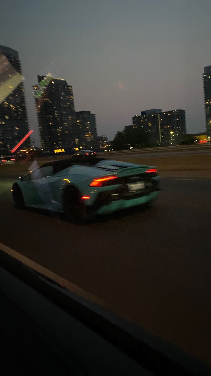 a blurry photo of a teal lamborghini driving on a highway at night. the photo is taken from inside a moving car, and the city skyline is visible in the background.
