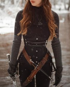 a woman stands in a snowy forest, dressed in a black corset with leather straps and a longsleeved top. she is armed with a sword and dagger, strapped to her waist. the woman has long brown hair and her gaze is directed offcamera. the image evokes a sense of fantasy and adventure, suggesting a character from a medieval or fantasy setting.