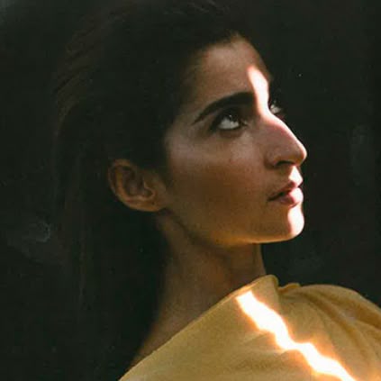 a closeup portrait of a woman looking up and to the right, with a strong ray of sunlight hitting her forehead and a diagonal ray across her shoulder. she is wearing a yellow garment. the background is dark, creating a strong contrast.