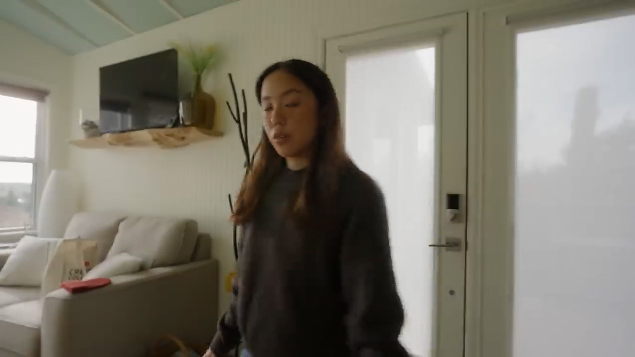 a young woman is standing in a living room and is facing the camera. she is wearing a dark grey sweater. the living room has a white couch with white pillows on it, a flatscreen tv mounted on the wall, and a glass door. there are some windows on the left side of the image.