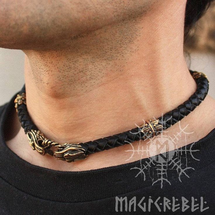 a closeup shot of a man wearing a braided leather necklace with bronze dragon head accents. the necklace is black and the bronze details have a vintage look. the man is wearing a black tshirt. a vegvisir symbol and the text "magicrebel" are visible in the lower part of the image, likely a watermark or brand logo. the focus is on the necklace and the man's neck area.