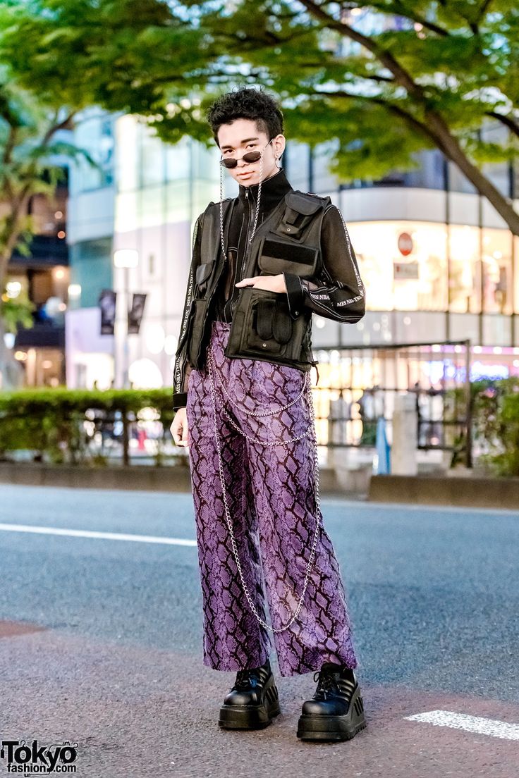 a full shot of a male model showcasing tokyo street style. he's wearing purple snakeskin print pants, a black vest, and chunky black boots.  the outfit is accessorized with multiple silver chains and sunglasses. the model is standing on a city street with blurred background buildings and trees. the overall aesthetic is edgy and contemporary.