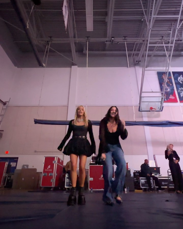 two women are dancing in a basketball court or similar large indoor space. one woman is blonde and wearing a black mini dress with chunky boots, while the other woman has long, dark hair and is wearing a black top and baggy jeans. in the background, there are stage crew members, equipment trucks, and a man with a microphone. a basketball hoop is visible on the right side of the image. the atmosphere is casual and energetic.