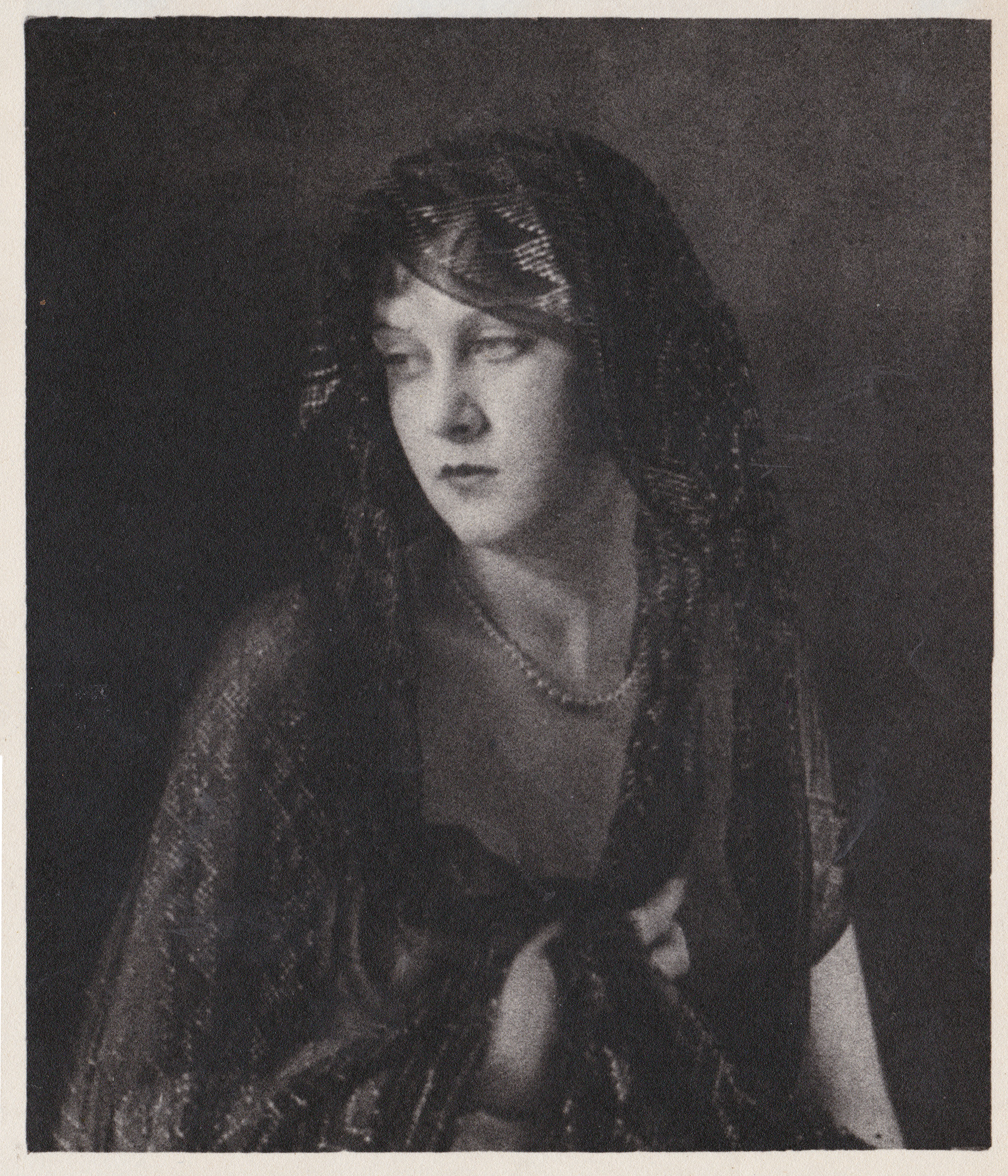a black and white portrait photograph of a young woman from the 1920s. she is wearing a dark, patterned dress or robe and a sheer dark veil over her head and shoulders. a pearl necklace is visible around her neck. her expression is serious and pensive. the background is dark and unfocused, drawing attention to the woman's features. the image has a vintage feel, typical of studio portraiture from the era.