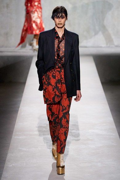 the image shows a fashion model walking down a runway. she is wearing a dark navy doublebreasted blazer over a red and black floralprinted pantsuit.  the pants are highwaisted and have a slightly cropped length. the model is also wearing gold platform shoes and has a short, dark bob hairstyle. the background is a muted grey, and a blurry figure wearing a red dress is visible in the background. the overall aesthetic suggests a fall/winter fashion collection.