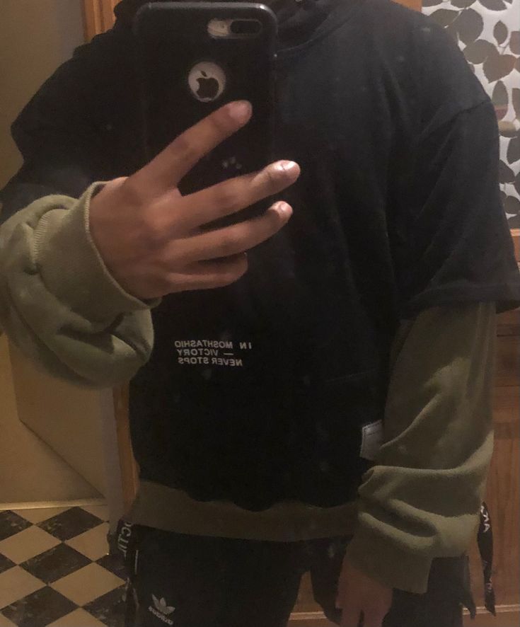 a man wearing a black tshirt over a longsleeved green hoodie. he is holding a black phone in his hand, with the screen facing the camera. there is writing on the tshirt. he is standing and looking down at the phone.