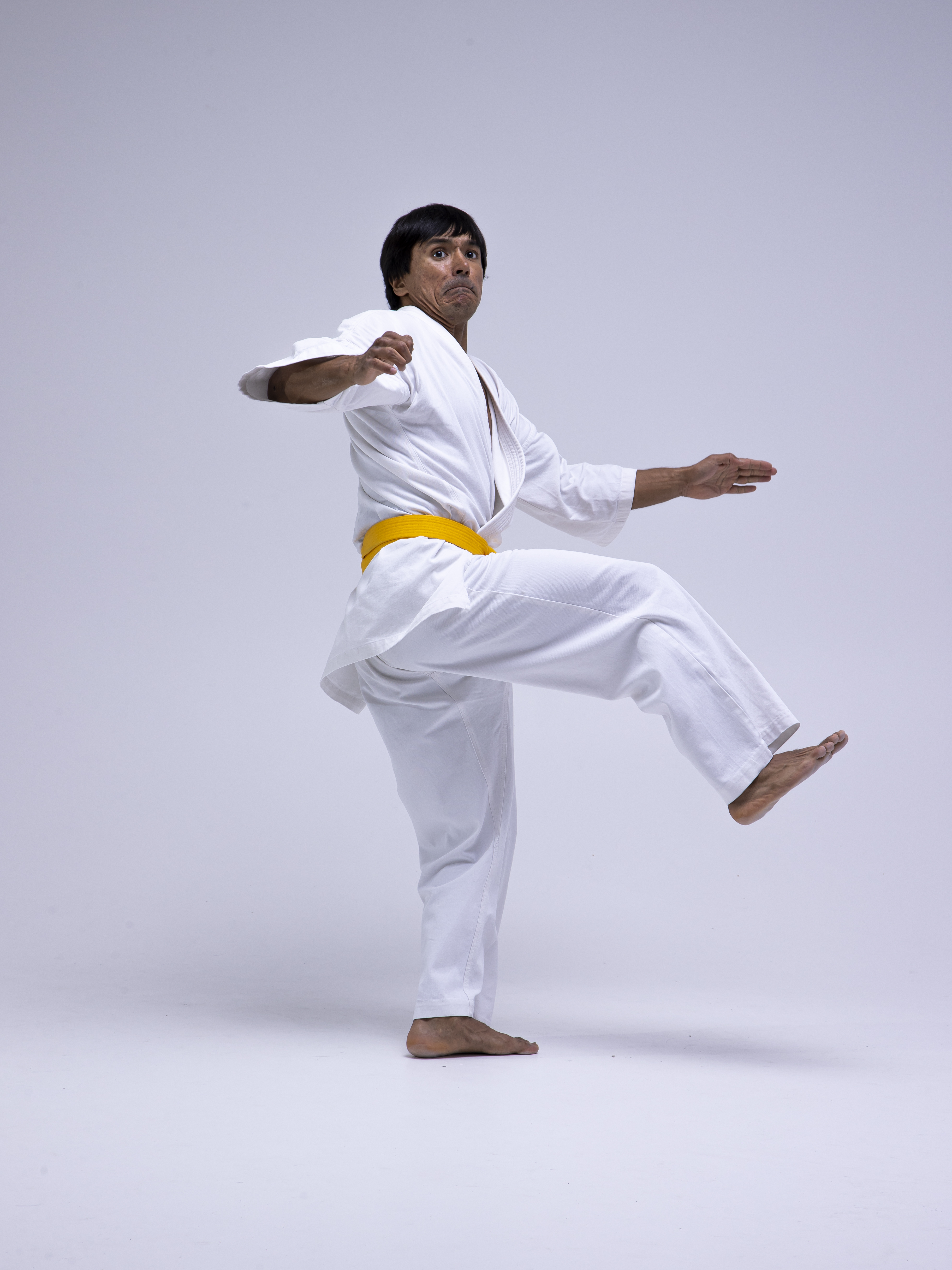 a man wearing a white karate gi with a yellow belt is performing a karate stance with one leg raised in a studio setting against a white background. he is barefoot and the image captures a full body shot of him in action.