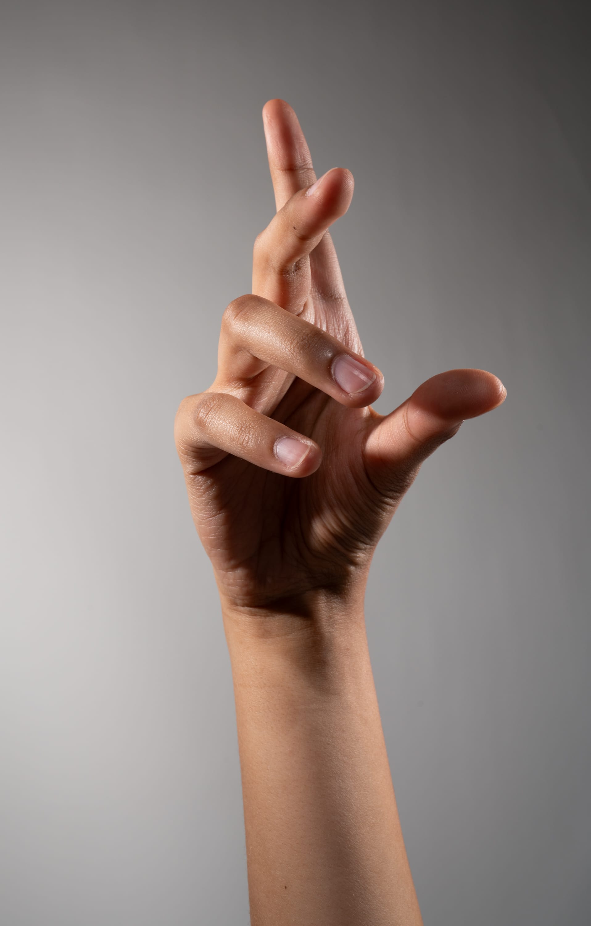 a hand with crossed fingers, gesturing hope or good luck. the hand is positioned against a neutral gray background, illuminated by soft studio lighting.