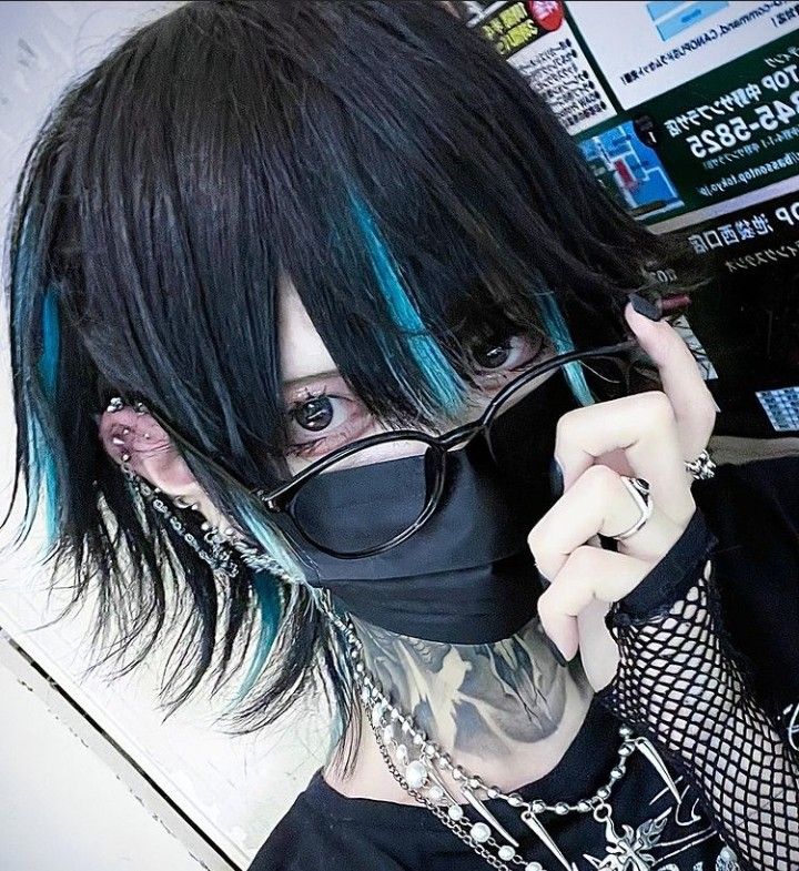 a closeup selfie of a person showcasing their unique style, leaning towards the visual kei japanese fashion subculture. they have dark blue hair with streaks of electric blue framing their face. they are wearing a black face mask, blackrimmed glasses, and multiple ear piercings. their accessories include layered necklaces with silver pendants and a black net glove on one hand. the background is slightly out of focus, hinting at an indoor setting. the lighting is artificial, possibly from a phone screen as they take the selfie.