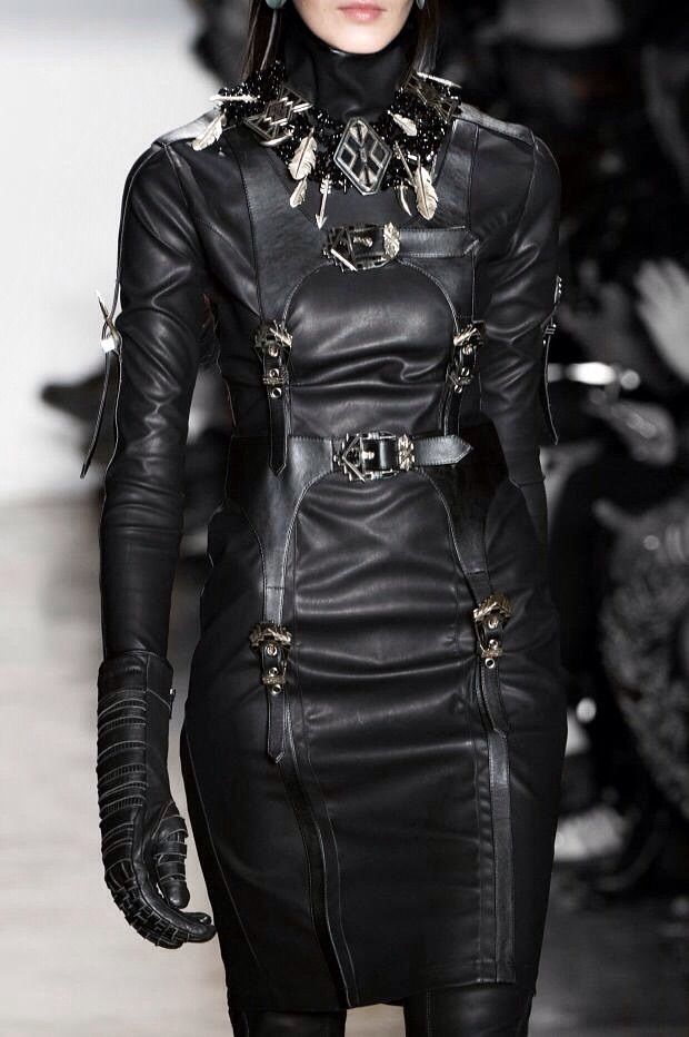 a woman is wearing a fitted black leather dress with long sleeves, kneelength, and a collar necklace with feathers and silver accents. the dress features belts and buckles. the woman also wears leather gloves. the image appears to be from a fashion show or runway, with a stage or catwalk in the background.