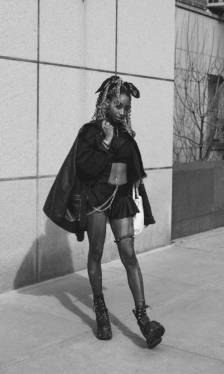 a black and white street style photograph of a young woman with braids walking down a city sidewalk. she is wearing a black jacket, a short black skirt, black boots, and jewelry. the background is a city building with a light colored wall. the overall style is edgy and alternative.