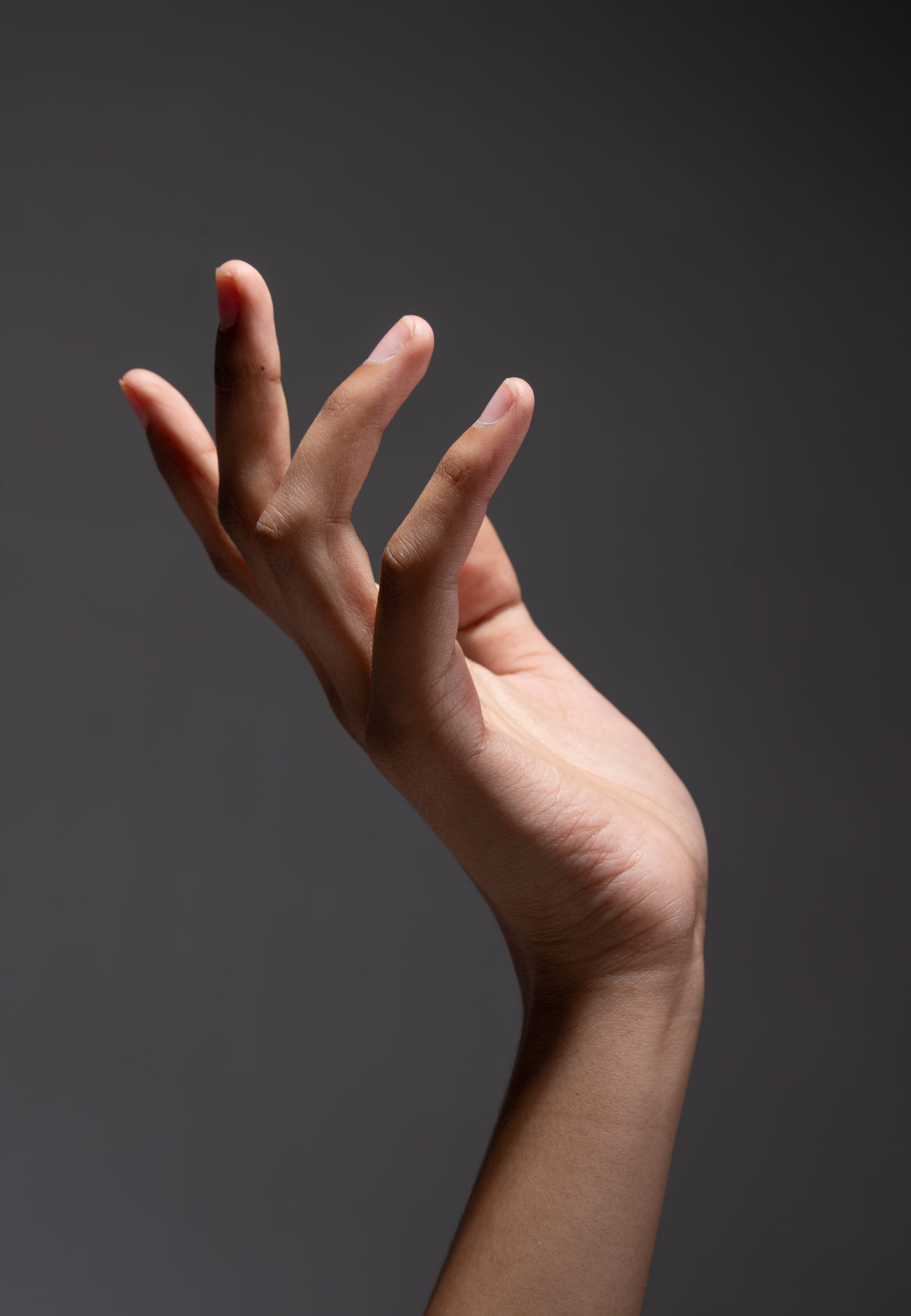 a graceful female hand is posed against a dark gray backdrop. the hand is delicately extended, with the fingers gently splayed. the studio lighting highlights the contours of the hand, showcasing its smooth skin and elegant pose.