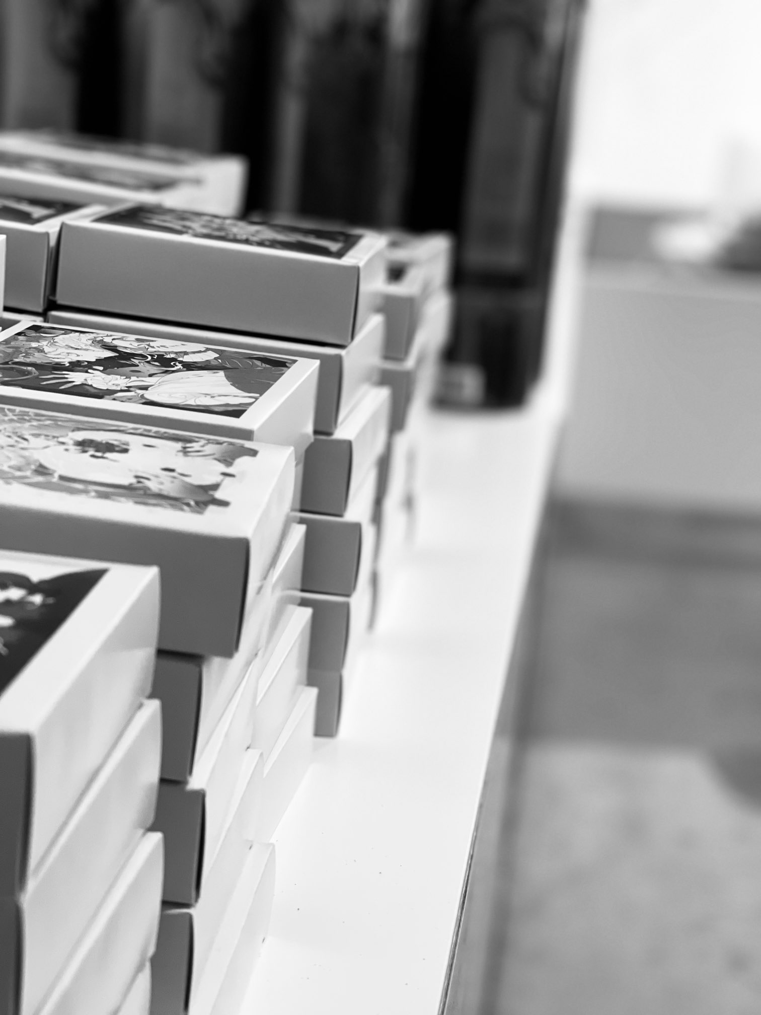 this is a black and white image of a stack of boxes on a shelf. the boxes are all the same size and shape, and they are arranged in a neat stack. the boxes appear to contain photographs. the image is taken from a low angle, so the viewer is looking up at the stack of boxes. the lighting is bright and even, and the image has a shallow depth of field.  the background is out of focus, but appears to be the corner of a room.