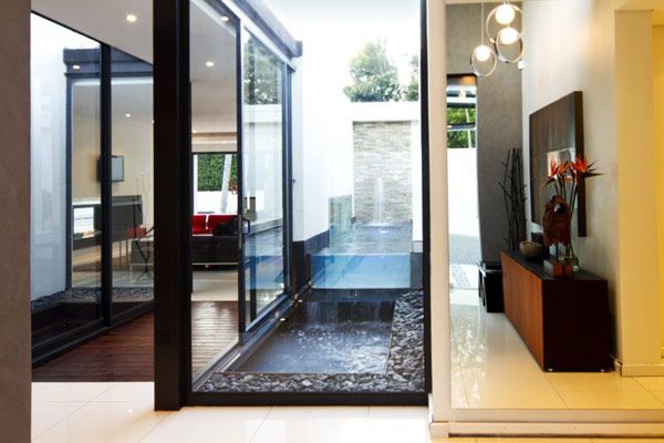 the image showcases a contemporary interior design, featuring a living room space with a unique water feature. sliding glass doors offer a view of an outdoor fountain area. the hallway is adorned with decor, plants, furniture, lighting fixtures, and artwork. the overall style is modern and sophisticated.