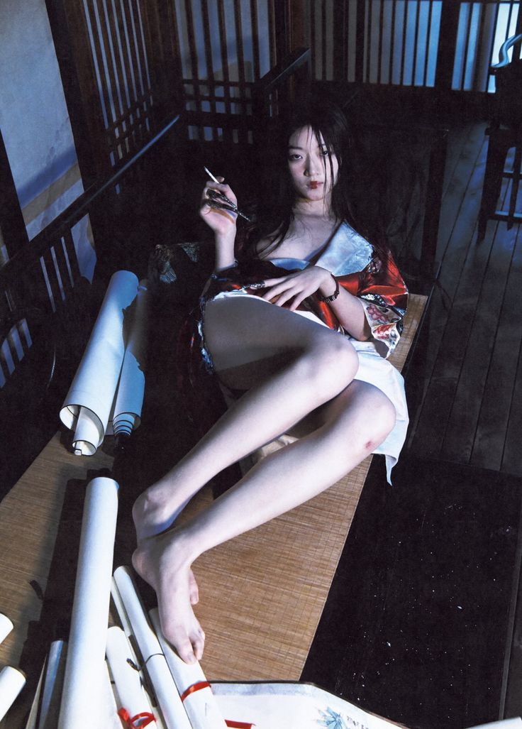 a woman is sitting on the floor indoors. she is wearing a kimono and holding a lit cigarette. several scrolls lie on the floor beside her.