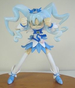 loseup view of an animestyle figurine of a girl, possibly a magical girl character. she's posed with her hands over her face in an expression of anger or frustration. her outfit is predominantly blue and white, typical of magical girl attire. the figurine is made of plastic and has detailed features, including hair, clothing, and boots. it's positioned on a wooden surface, suggesting it is a collectible item. the background is a simple, lightcolored surface that doesn't distract from the figure. the overall style and design point towards it being a collectible toy derived from an anime or manga series.