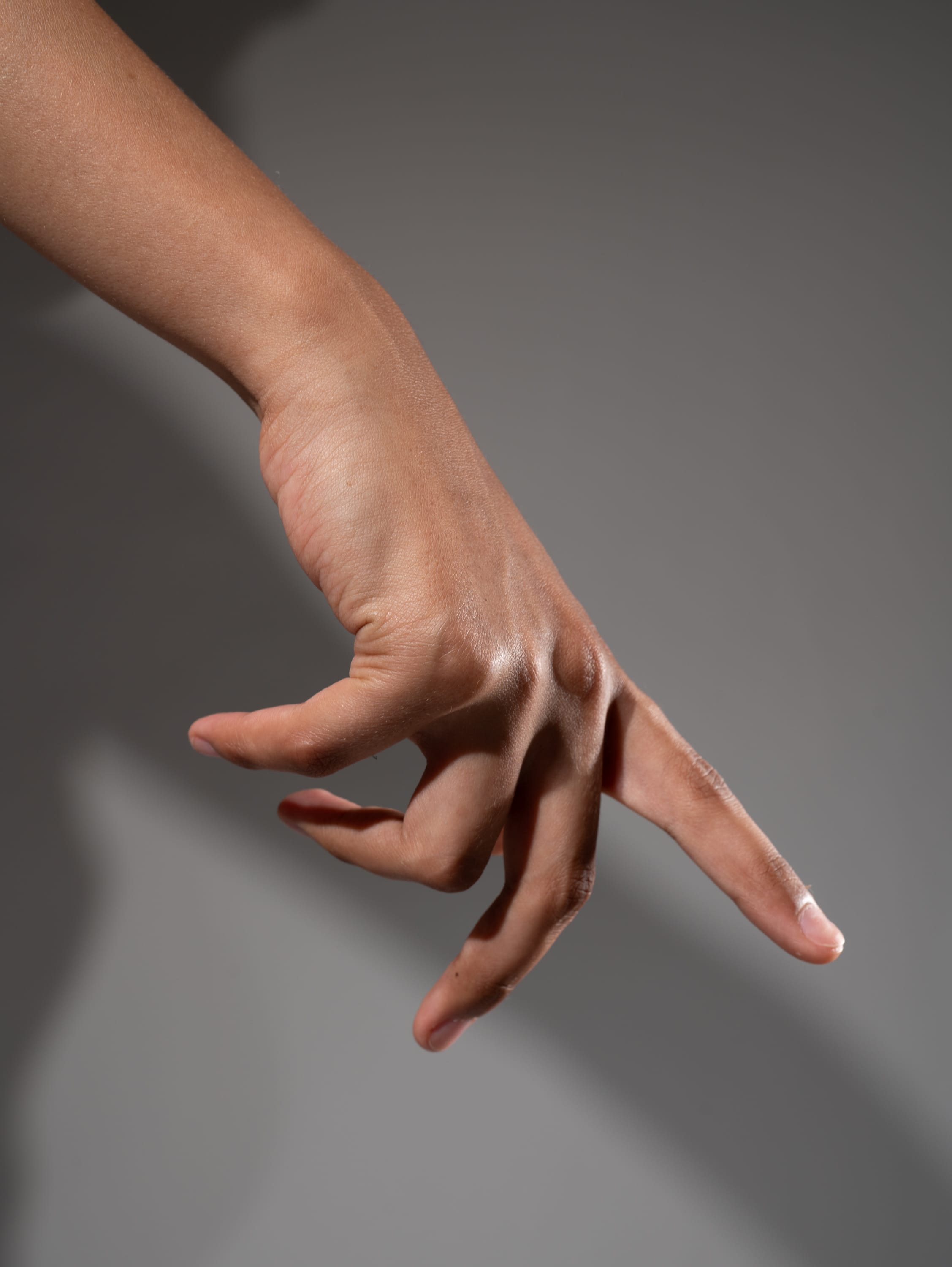 a closeup image of a hand with light brown skin reaching down with the index finger outstretched, as if pointing or reaching for something below. the lighting is soft and creates a subtle shadow on the hand and the background, highlighting the contours and texture of the skin. the background is a plain grey, allowing the focus to remain solely on the hand and its gesture.