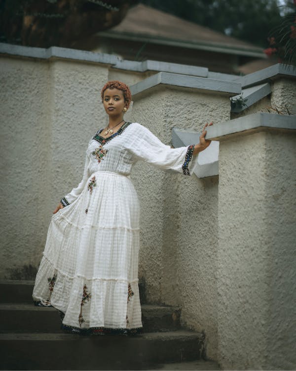 a woman in a white embroidered dress and jewelry is standing on a set of steps next to a textured white wall. the steps lead upwards, and a building is visible in the background. she is holding onto the top edge of the wall. the overall style of the image suggests a cultural or traditional setting.