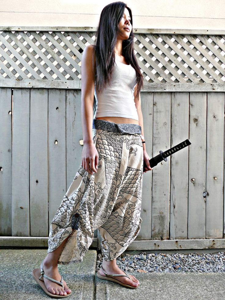 a young asian woman dressed in a samuraiinspired outfit.  she's wearing a white tank top, cream hakama pants with a black and white design, and black sandals. she's holding a katana (samurai sword) in her right hand. she's standing in front of a wood fence with a lattice design.