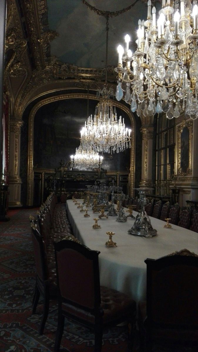the image shows a lavishly decorated dining room, possibly in a palace or mansion. the room is dimly lit, with the main light sources being several large, ornate crystal chandeliers hanging from a high, gilded ceiling. the walls are dark and richly decorated with gold detailing and what appears to be dark artwork or tapestries. a long, formal dining table is set with silverware, and a number of dark, upholstered chairs are arranged around it.  the overall atmosphere is one of opulence, elegance, and historical grandeur. the image is evocative of a bygone era of aristocratic wealth and sophistication.