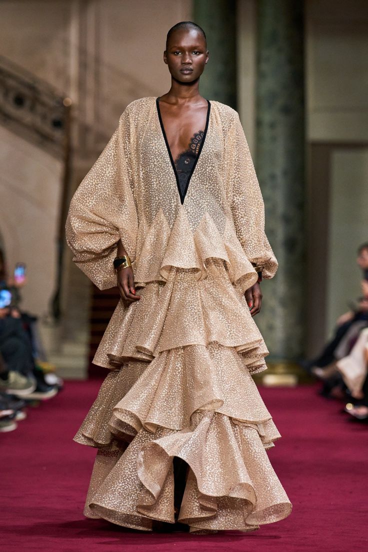 a darkskinned model walks down a runway in a stunning, gold, glittery gown. the dress features layers of ruffles, and a plunging neckline. she is wearing a black, lace bodysuit underneath the dress. the model has short black hair and is wearing minimal makeup. she is carrying a black clutch. the background is a blur of the runway and other models.