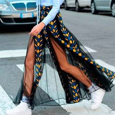 a woman wearing a black and blue floral print skirt is walking down the street. she has a white shirt on and is wearing white sneakers. the skirt has a slit up the side and is a sheer material. the woman is walking on a sidewalk and there are cars parked on the street in the background.