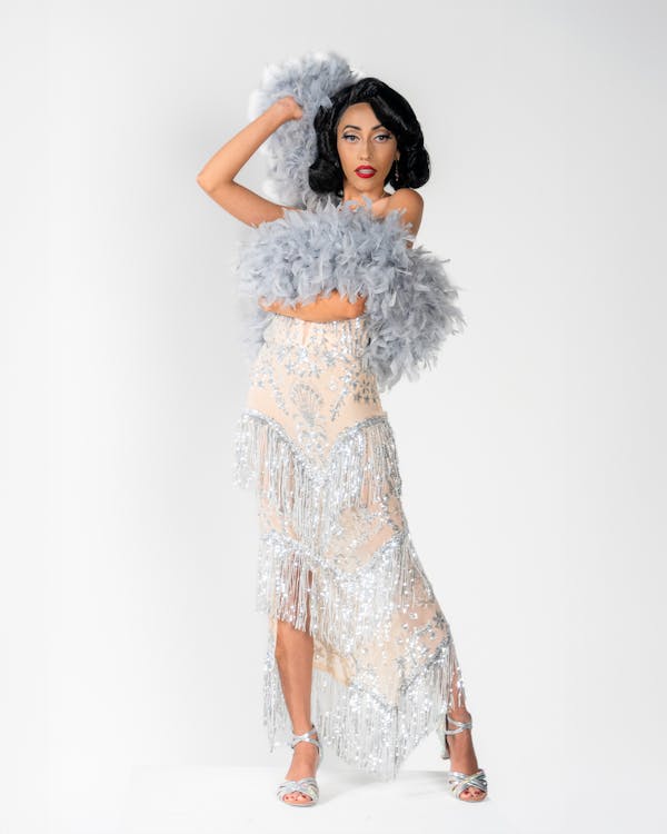 a full shot of a woman standing in front of a white background. she is wearing a 1920s showgirl style dress with silver fringe and a feather boa around her neck. she is wearing silver heels.